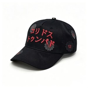 May include: Black baseball cap with red Japanese text and flower patches. The cap has embroidered text and several flower-shaped patches in grey and red. A stylish accessory for casual wear.