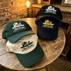 May include: A collection of three baseball caps featuring the Aim&eacute; Leon Dore logo and globe graphic. One cap is dark green, one is black with white embroidery, and one is navy blue with yellow embroidery. A cream cap with a dark green brim is included.