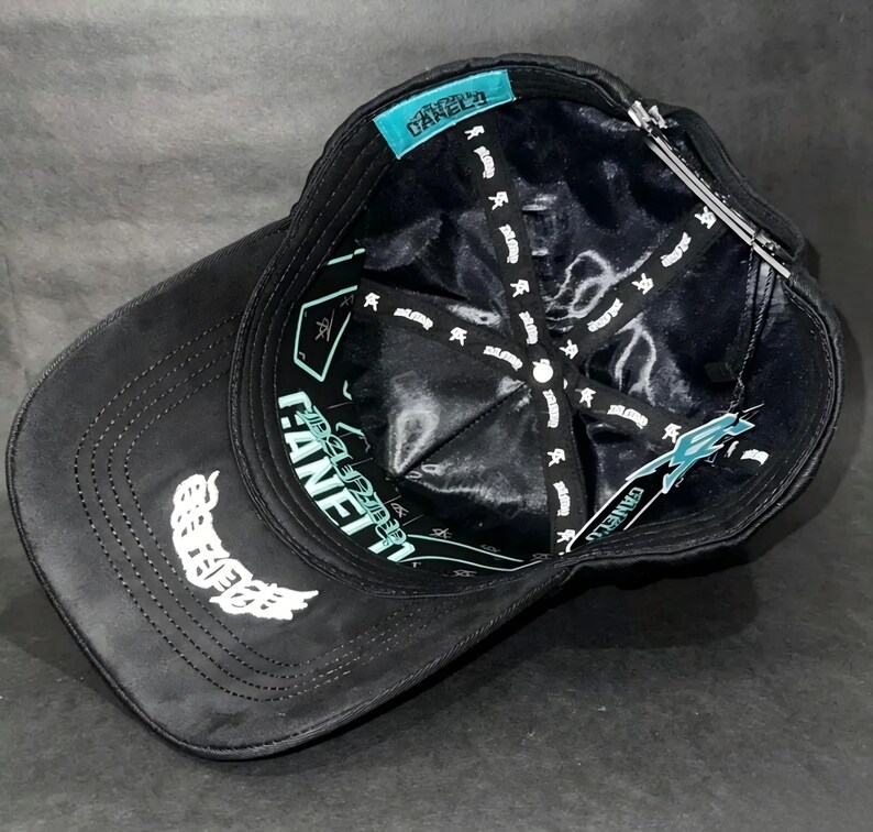 May include: Black baseball cap with white embroidered wing design on the side. The interior features a black lining with white text and a teal tag with the brand name. The cap has a curved brim.