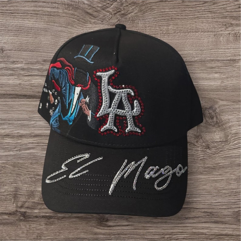May include: Black baseball cap with the letters "LA" in silver rhinestones. The cap features an embroidered design of a magician and the words "El Mago" in silver script on the brim.