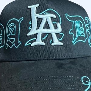 K&ouml;nnte beinhalten: Schwarze Baseballkappe mit dem gestickten Text "LA Dad" in Hellblau. Die Kappe hat einen gebogenen Schirm und eine strukturierte Krone, wobei der Text in einer stilisierten Schriftart gehalten ist.