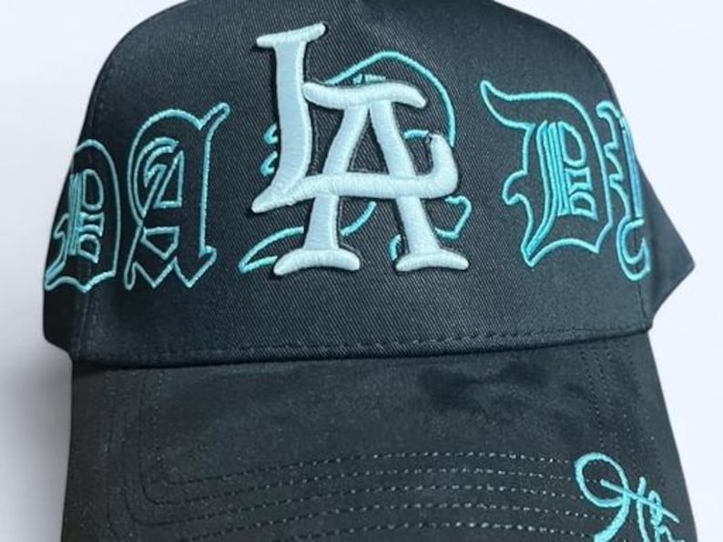 K&ouml;nnte beinhalten: Schwarze Baseballkappe mit dem gestickten Text "LA Dad" in Hellblau. Die Kappe hat einen gebogenen Schirm und eine strukturierte Krone, wobei der Text in einer stilisierten Schriftart gehalten ist.