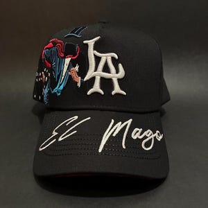May include: Black baseball cap with white embroidered text "LA" and "El Mago". The cap features a colorful embroidered design of a figure in a cape. The brim is curved and the cap is made of a textured fabric.