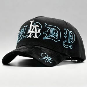 K&ouml;nnte beinhalten: Schwarze Baseballkappe mit den Buchstaben "LA" in Wei&szlig; und "D" in hellblauer Stickerei. Die Kappe hat einen gebogenen Schirm und eine strukturierte Krone. Der Text "9th" ist auf der Seite gestickt.