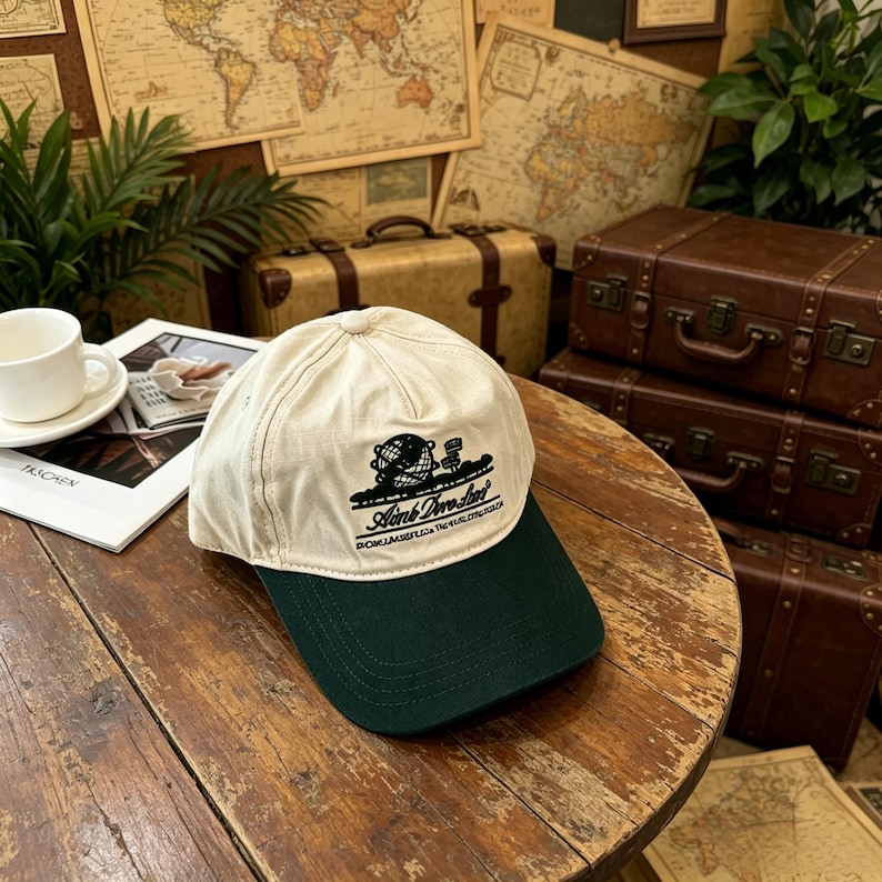 May include: A two-tone baseball cap featuring an off-white crown and a dark green brim. The front panel displays a black embroidered graphic of a globe with a small flag and the text "Aimb Dwo Sun" with smaller text underneath.
