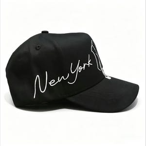 May include: Black baseball cap with white embroidered text that reads "New York". The cap has a curved brim and a small button on top. The design includes a white outline of the Statue of Liberty.