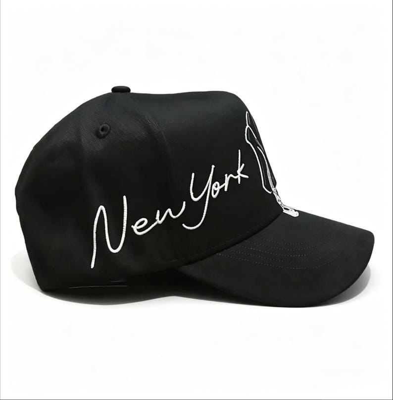 May include: Black baseball cap with white embroidered text that reads "New York". The cap has a curved brim and a small button on top. The design includes a white outline of the Statue of Liberty.