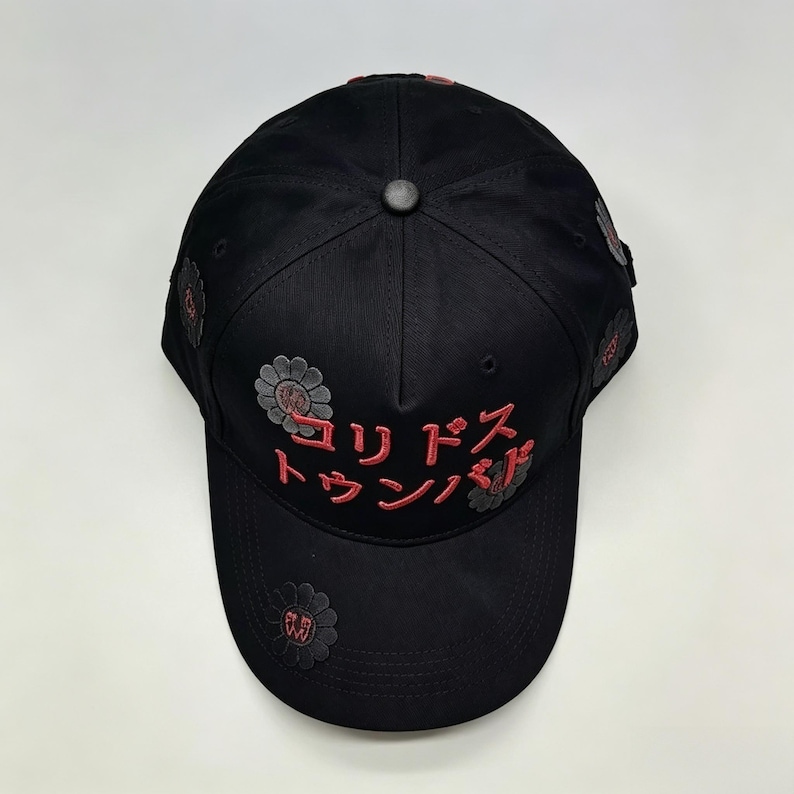May include: Black baseball cap with red Japanese text and flower designs. The cap has a curved peak and a button on top. The Japanese text is embroidered in red, and the flower designs are grey and red.