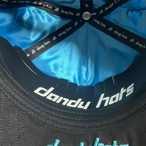 K&ouml;nnte beinhalten: Schwarze Baseballkappe mit t&uuml;rkisblauem Satinfutter. Die Unterseite des Schirms tr&auml;gt den wei&szlig;en Schriftzug "dandy hats". Die Kappe hat ein schwarzes Band mit dem Text "ch dandy here".