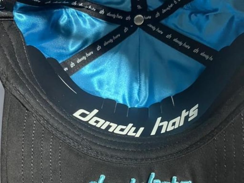K&ouml;nnte beinhalten: Schwarze Baseballkappe mit t&uuml;rkisblauem Satinfutter. Die Unterseite des Schirms tr&auml;gt den wei&szlig;en Schriftzug "dandy hats". Die Kappe hat ein schwarzes Band mit dem Text "ch dandy here".