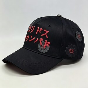 May include: Black baseball cap with red Japanese text and flower designs. The cap features embroidered text on the front and several flower patches. The cap has a curved peak and a button on top.