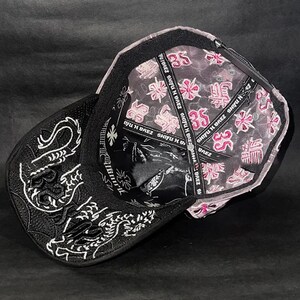 Peut inclure: Casquette de baseball noire avec broderie de dragon blanc sur le c&ocirc;t&eacute;. L'int&eacute;rieur pr&eacute;sente un motif floral rose et blanc et le texte "SHIFU H BREZ". La casquette a une visi&egrave;re incurv&eacute;e.