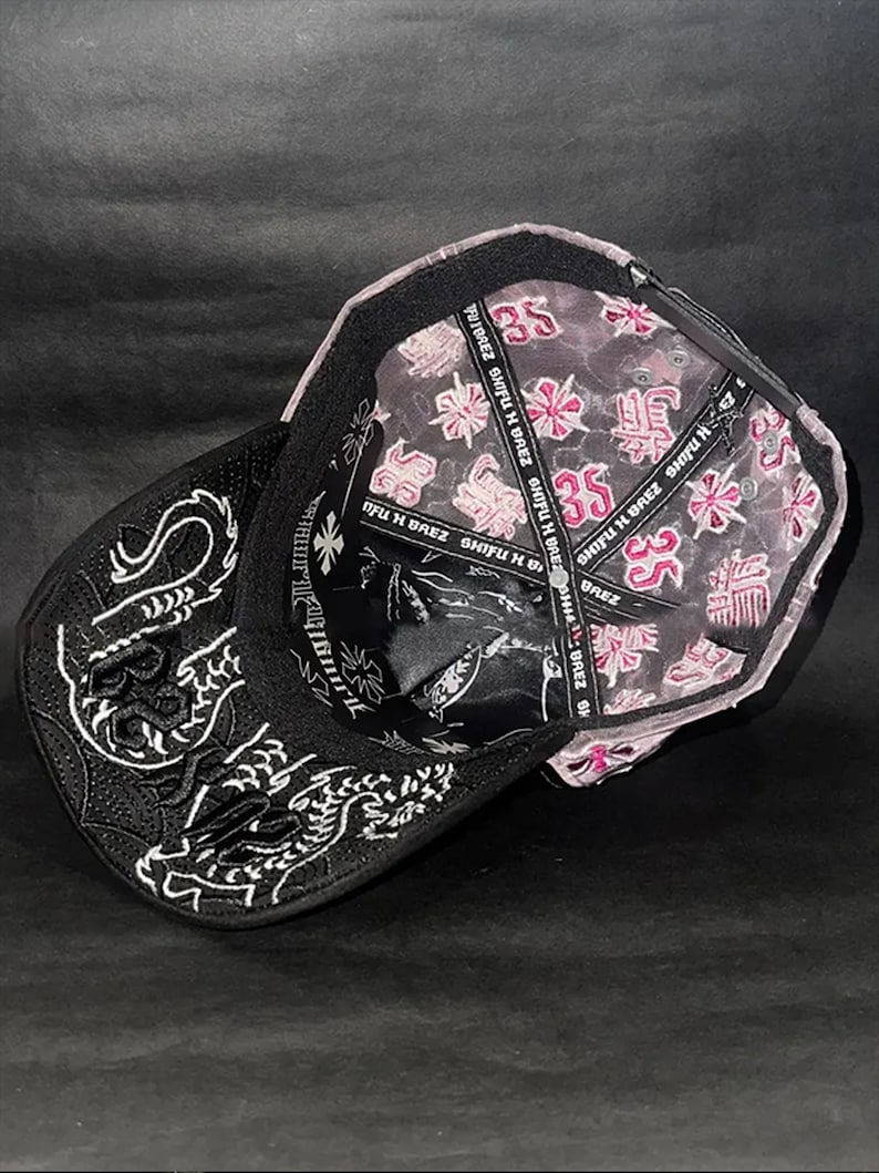 Peut inclure: Casquette de baseball noire avec broderie de dragon blanc sur le c&ocirc;t&eacute;. L'int&eacute;rieur pr&eacute;sente un motif floral rose et blanc et le texte "SHIFU H BREZ". La casquette a une visi&egrave;re incurv&eacute;e.