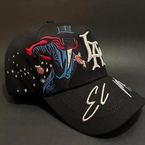 May include: Black baseball cap with embroidered design. Features a magician in a blue suit and red cape, with the letters "LA" in white. The brim has the text "EL 13" in white script. The cap has a curved brim.