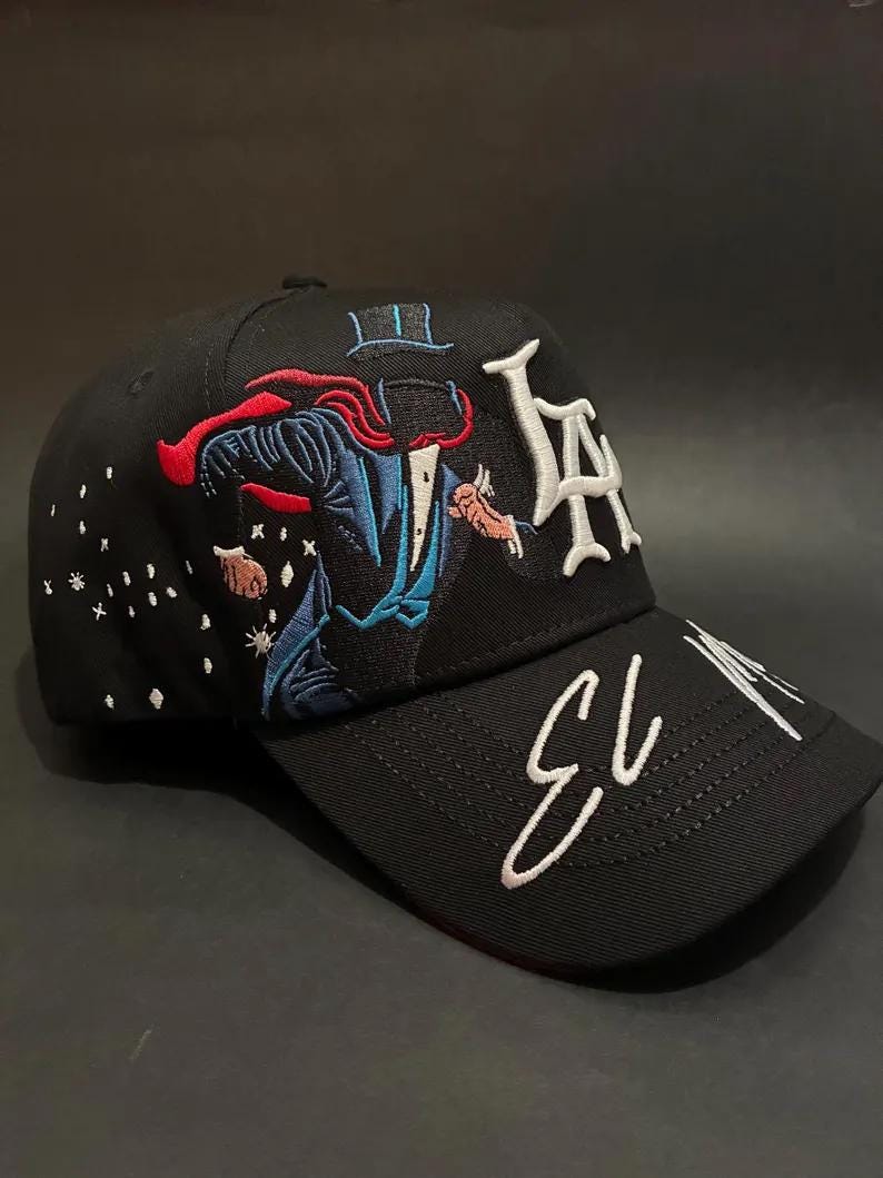 May include: Black baseball cap with embroidered design. Features a magician in a blue suit and red cape, with the letters "LA" in white. The brim has the text "EL 13" in white script. The cap has a curved brim.