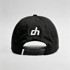 K&ouml;nnte beinhalten: Schwarze Baseballkappe mit wei&szlig;em "dh"-Logo auf der Vorderseite. Der verstellbare Riemen tr&auml;gt den Schriftzug "dune hats" in einer stilisierten Schriftart. Die Kappe ist aus strapazierf&auml;higem Stoff gefertigt und hat einen gebogenen Schirm.