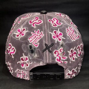 Peut inclure: Casquette de baseball grise avec une visi&egrave;re noire et une sangle r&eacute;glable. La casquette pr&eacute;sente des motifs floraux et des caract&egrave;res chinois roses brod&eacute;s. Les lettres "B", "X" et "A" sont &eacute;galement visibles.