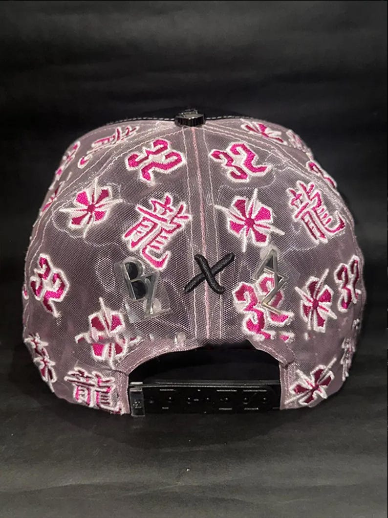 Peut inclure: Casquette de baseball grise avec une visi&egrave;re noire et une sangle r&eacute;glable. La casquette pr&eacute;sente des motifs floraux et des caract&egrave;res chinois roses brod&eacute;s. Les lettres "B", "X" et "A" sont &eacute;galement visibles.