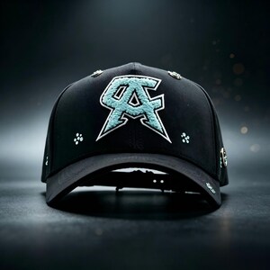 May include: Black baseball cap with a teal and white embroidered logo. The cap features a curved brim and decorative studs around the crown. The logo is a stylized "AC" design.
