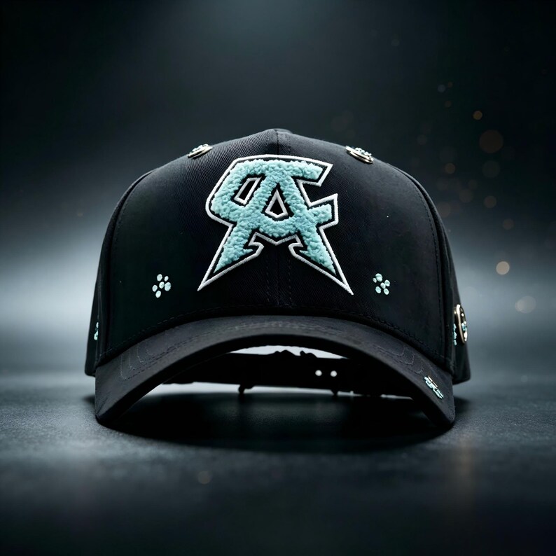 May include: Black baseball cap with a teal and white embroidered logo. The cap features a curved brim and decorative studs around the crown. The logo is a stylized "AC" design.