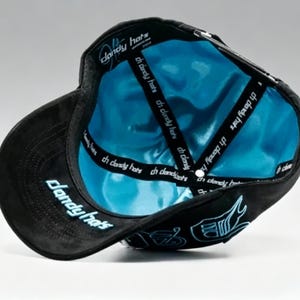K&ouml;nnte beinhalten: Schwarze Baseballkappe mit hellblauem Satin-Innenfutter. Die Kappe hat den Text "dandy hats" in Hellblau auf der Krempe und Seite. Im Inneren befindet sich der Text "ch dandy hats" auf den Riemen.