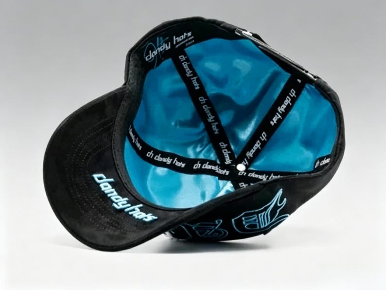 K&ouml;nnte beinhalten: Schwarze Baseballkappe mit hellblauem Satin-Innenfutter. Die Kappe hat den Text "dandy hats" in Hellblau auf der Krempe und Seite. Im Inneren befindet sich der Text "ch dandy hats" auf den Riemen.
