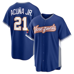 Puede incluir: Una camiseta de béisbol azul con el nombre "ACUÑA JR." y el número "21" en la espalda. La parte delantera de la camiseta muestra la palabra "Venezuela" en una fuente cursiva. La camiseta tiene detalles en blanco y naranja.