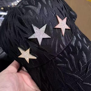 May include: Black baseball cap with a textured leaf pattern and three star embellishments. The stars are silver, rose gold, and gold, each with a sparkling, glittery appearance. The cap is held by a hand.