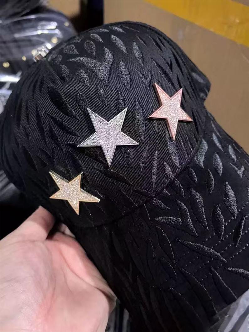 May include: Black baseball cap with a textured leaf pattern and three star embellishments. The stars are silver, rose gold, and gold, each with a sparkling, glittery appearance. The cap is held by a hand.