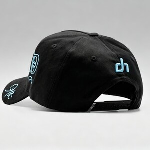 K&ouml;nnte beinhalten: Schwarze Baseballkappe mit flachem Schirm und verstellbarem Snapback-Verschluss. Die Kappe hat hellblaue gestickte Designs auf der Vorderseite, Seite und R&uuml;ckseite. Klassischer, sportlicher Stil.