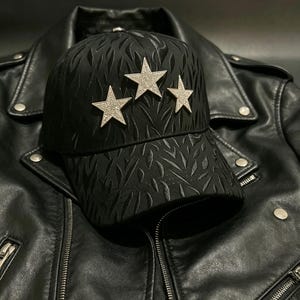 May include: Black baseball cap with three silver star embellishments. The cap has a textured pattern and is set against a black leather jacket. The jacket features a zipper closure and silver hardware.