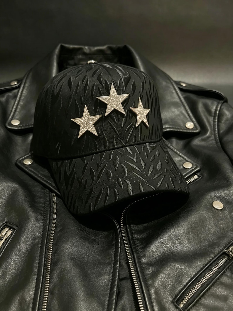 May include: Black baseball cap with three silver star embellishments. The cap has a textured pattern and is set against a black leather jacket. The jacket features a zipper closure and silver hardware.