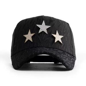 May include: Black baseball cap with a textured pattern and three star embellishments. The stars are silver and gold, adding a touch of sparkle to the cap. This stylish cap is a fashion accessory.