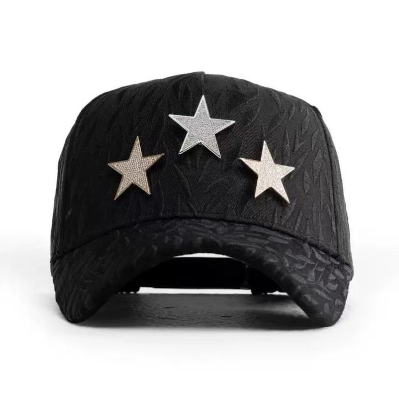 May include: Black baseball cap with a textured pattern and three star embellishments. The stars are silver and gold, adding a touch of sparkle to the cap. This stylish cap is a fashion accessory.