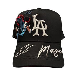 May include: Black baseball cap with white embroidered "LA" letters and "El Mago" script. The cap features colorful embroidery of a figure in a cape. A stylish accessory for casual wear.