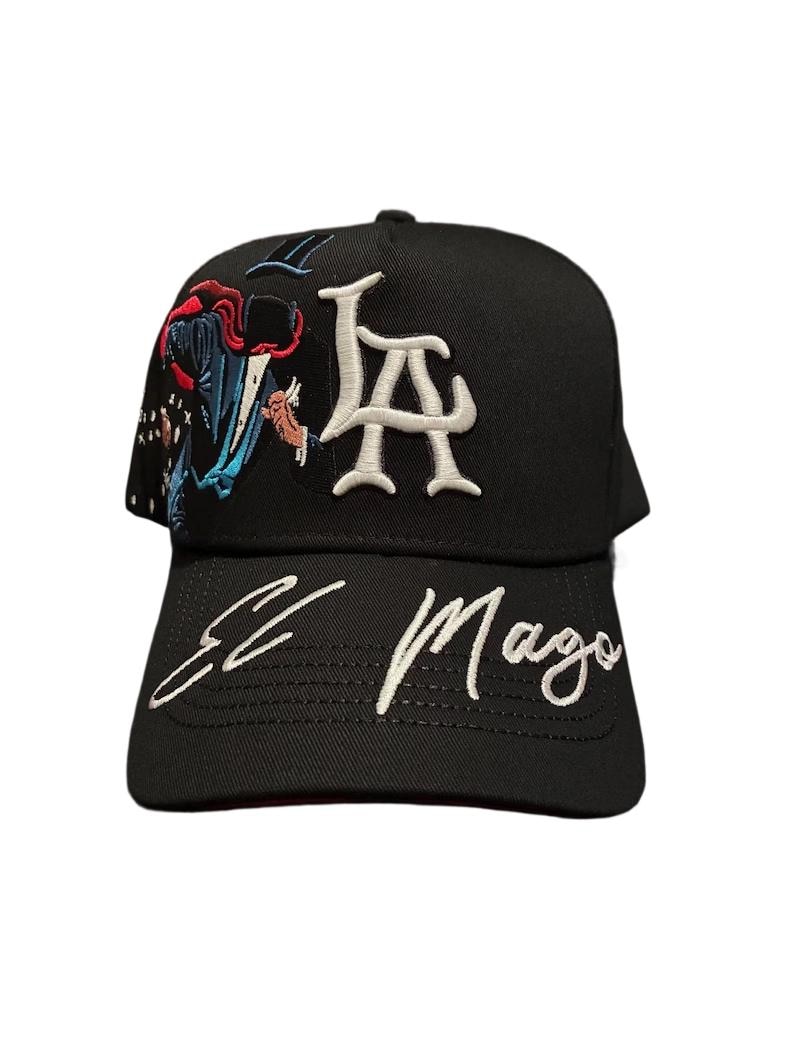 May include: Black baseball cap with white embroidered "LA" letters and "El Mago" script. The cap features colorful embroidery of a figure in a cape. A stylish accessory for casual wear.