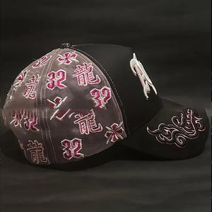 Peut inclure: Casquette de baseball noire avec des d&eacute;tails brod&eacute;s blancs et roses. La casquette pr&eacute;sente la lettre "R" en blanc sur le devant, et des textes et motifs floraux roses sur le c&ocirc;t&eacute; et &agrave; l'arri&egrave;re. Le panneau arri&egrave;re est en mat&eacute;riau gris transparent.
