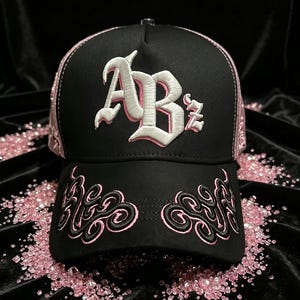 Peut inclure: Casquette de baseball noire avec des d&eacute;tails et broderies roses. La casquette pr&eacute;sente les lettres "ABz" en blanc avec un contour rose sur le devant. Des volutes roses brod&eacute;es ornent la visi&egrave;re. Le design de la casquette est pos&eacute; sur un fond noir.