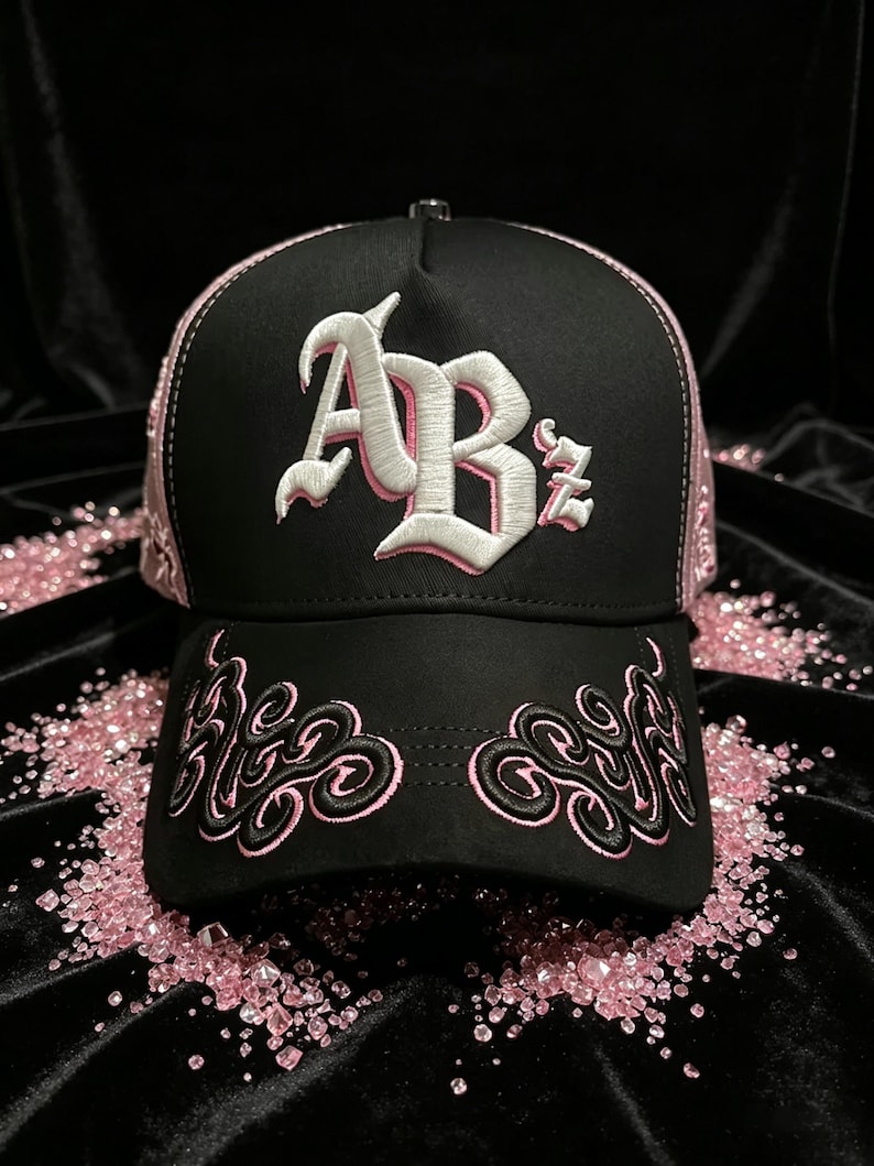 Peut inclure: Casquette de baseball noire avec des d&eacute;tails et broderies roses. La casquette pr&eacute;sente les lettres "ABz" en blanc avec un contour rose sur le devant. Des volutes roses brod&eacute;es ornent la visi&egrave;re. Le design de la casquette est pos&eacute; sur un fond noir.