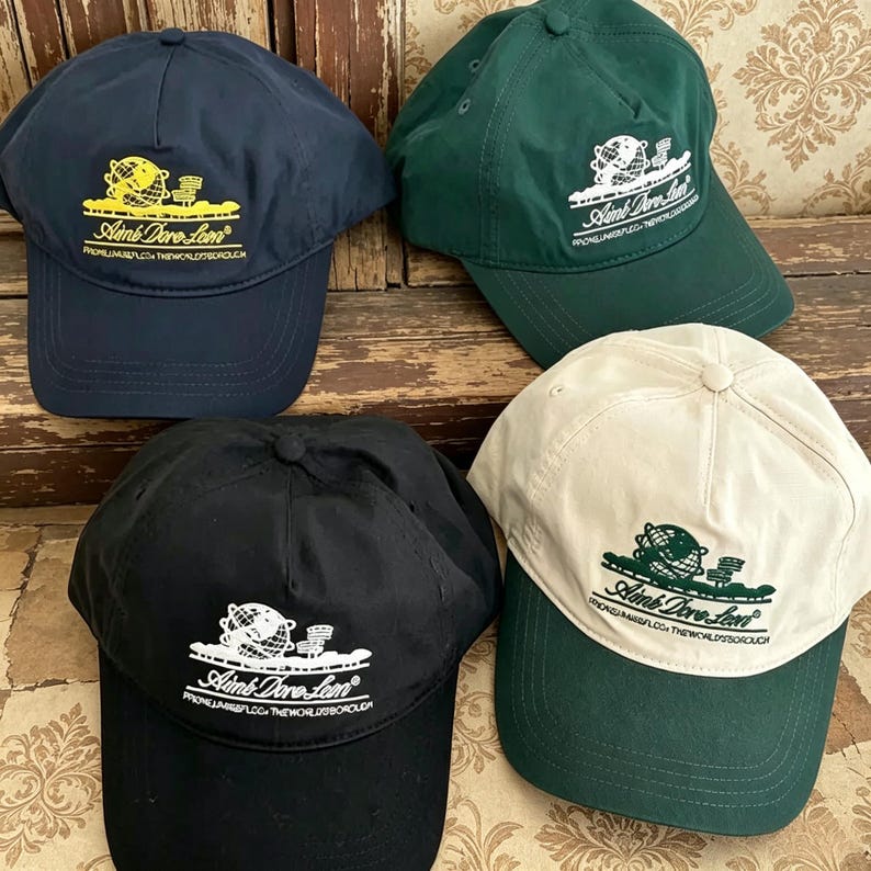 May include: Four baseball caps in navy blue, dark green, black, and cream. Each cap features an embroidered graphic of a globe and the text Aim&eacute; Leon Dore, along with the text Queens, NY, USA, The World's Borough printed below the logo.