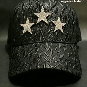 May include: Black baseball cap with a textured pattern and three silver star embellishments. The stars are encrusted with small, sparkling crystals. The cap has a curved brim and a structured design.