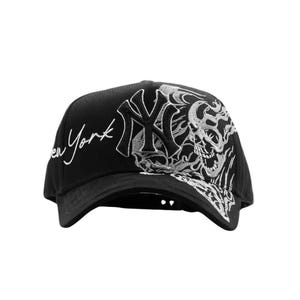 May include: Black baseball cap featuring a white embroidered skull design and the text "en York". The cap also displays the New York Yankees logo in white. The cap has a curved brim and a structured crown.