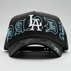 K&ouml;nnte beinhalten: Schwarze Baseballkappe mit dem Text "DAD" in einer stilisierten Schriftart, mit "LA" in Wei&szlig;. Der Text ist hellblau umrandet. Die Kappe hat einen gebogenen Schirm und eine Signatur auf der Seite.