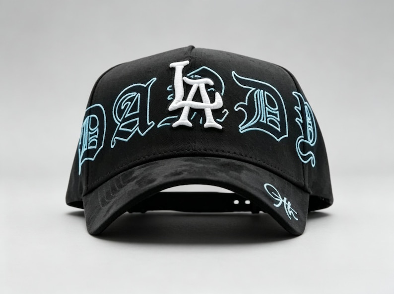 K&ouml;nnte beinhalten: Schwarze Baseballkappe mit dem Text "DAD" in einer stilisierten Schriftart, mit "LA" in Wei&szlig;. Der Text ist hellblau umrandet. Die Kappe hat einen gebogenen Schirm und eine Signatur auf der Seite.