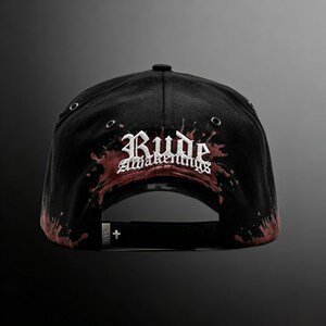 May include: Black baseball cap with the text "Rude Awakenings" in white gothic lettering, accented with red splatter design. The cap has a curved brim, adjustable strap, and metal eyelets.