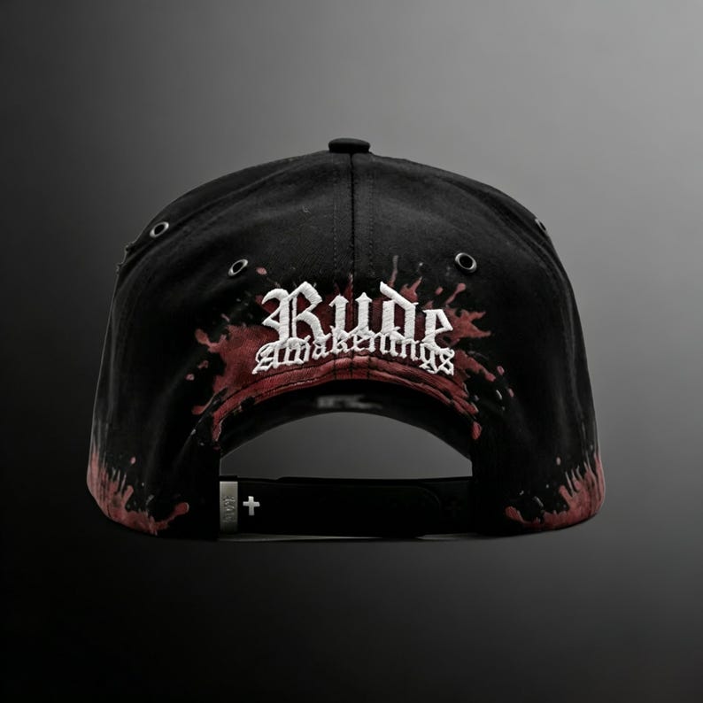 May include: Black baseball cap with the text "Rude Awakenings" in white gothic lettering, accented with red splatter design. The cap has a curved brim, adjustable strap, and metal eyelets.