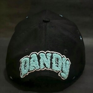 May include: Black baseball cap with the word "DANDY" embroidered in a light blue, fuzzy font. The cap has a curved brim and a button on top. The text is outlined in white.