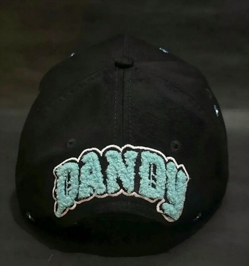 May include: Black baseball cap with the word "DANDY" embroidered in a light blue, fuzzy font. The cap has a curved brim and a button on top. The text is outlined in white.