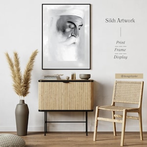 May include: A black framed print of Sikh artwork depicting a grayscale portrait of a man with a white turban and beard. The print hangs above a wooden cabinet with a vase of dried plants and a woven chair.