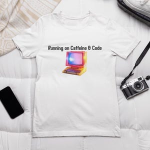 Puede incluir: Camiseta blanca con la frase "Running on Caffeine & Code" y un gráfico de ordenador retro colorido. El ordenador tiene una pantalla con tonos rosas, azules y amarillos. También se ven un smartphone negro y una cámara vintage.